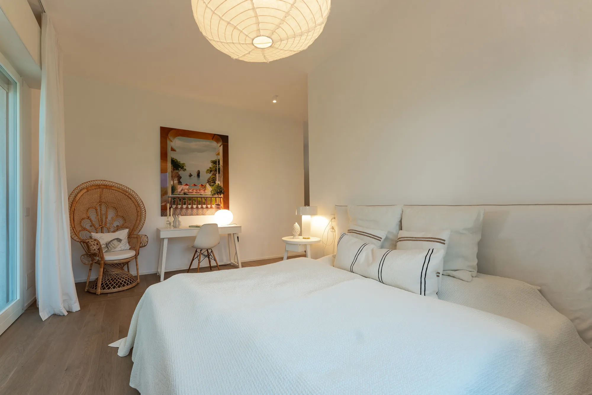 Bright bedroom with a white bed, a rattan peacock chair and a painting of the Riviera
