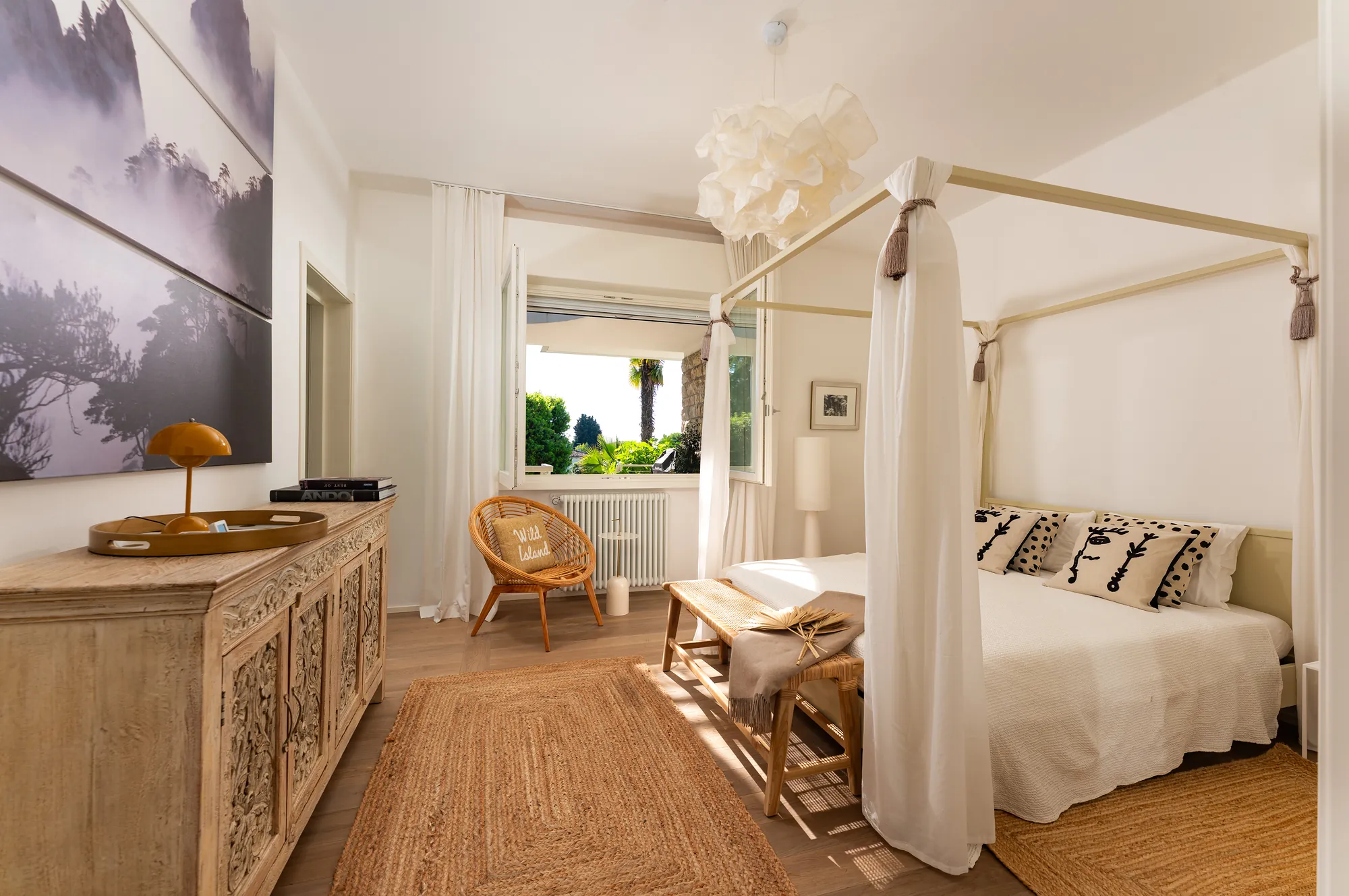 White four-poster bedroom with linen curtains, carved sideboard and garden view