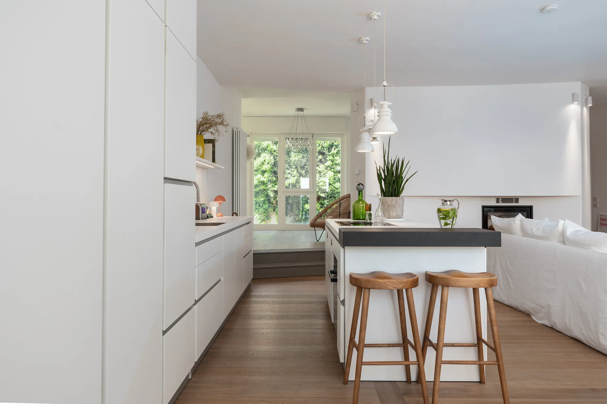 White kitchen with island and wooden bar stools, open to the living room with white sofas