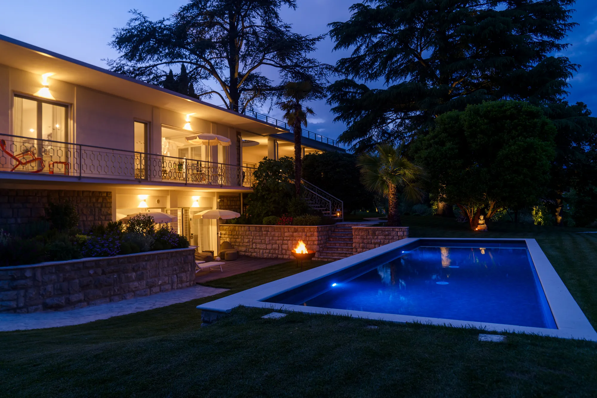 The pool glowing blue at night in front of the lit-up villa and its stone terraces