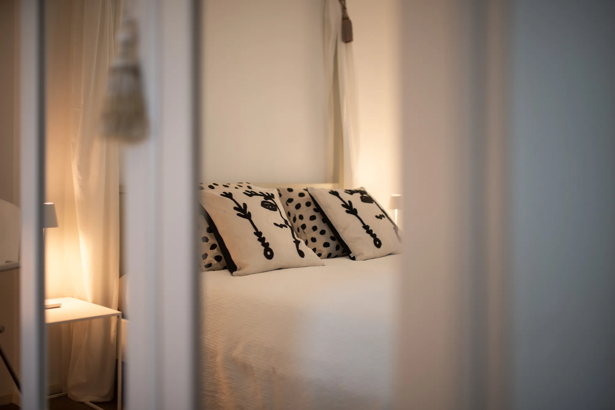 Black-and-white patterned cushions behind sheer bed curtains in soft lamplight